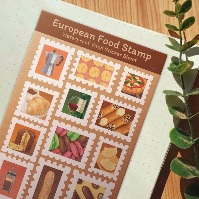 A waterproof vinyl sticker sheet featuring European food illustrations, styled on a light-colored notebook next to a green plant on a wooden table.