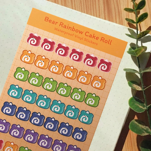 A sheet of colorful bear-shaped vinyl stickers labeled 'Bear Rainbow Cake Roll Waterproof Vinyl Stickers' placed on a white dotted notebook, with a wooden surface and green plant leaves visible around it.