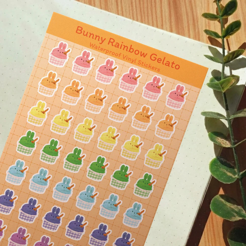 A sheet of 'Bunny Rainbow Gelato' waterproof vinyl stickers displayed on a notebook next to a green plant leaf. The stickers feature cute bunnies in pastel rainbow colors inside pink, yellow, green, blue, and purple gelato cups.