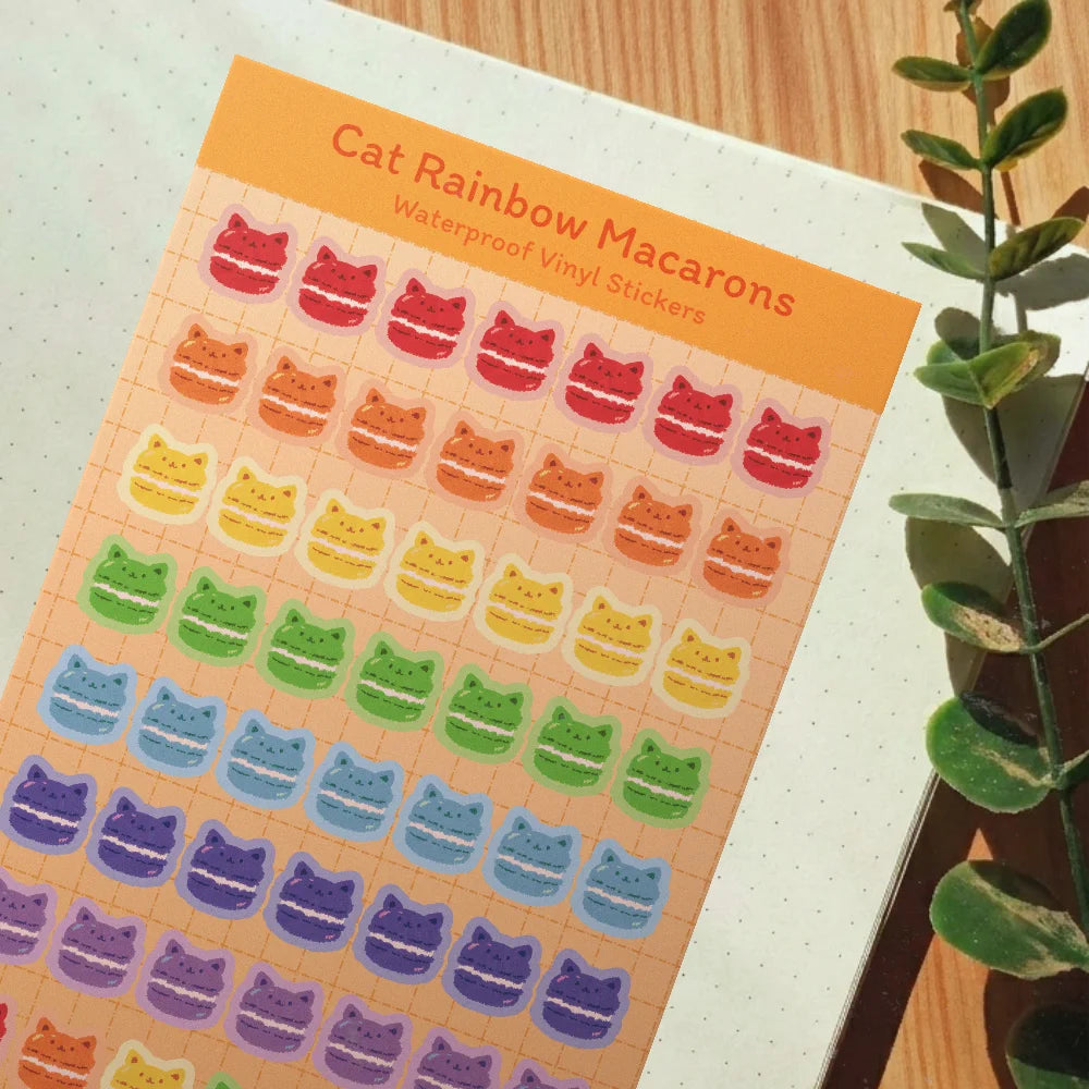 A sheet of cat-shaped rainbow macaron vinyl stickers displayed on a notebook page, with a green plant leaf visible on the right side.