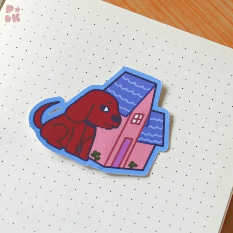 A die-cut sticker featuring a red dog sitting in front of a pink house with a blue wavy roof, placed on a white pegboard notebook.
