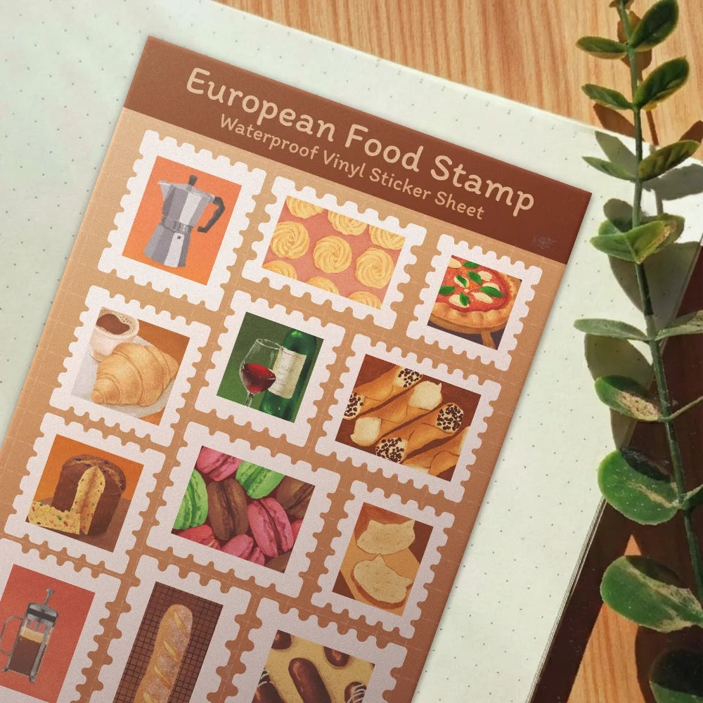 A waterproof vinyl sticker sheet featuring European food illustrations, styled on a light-colored notebook next to a green plant on a wooden table.